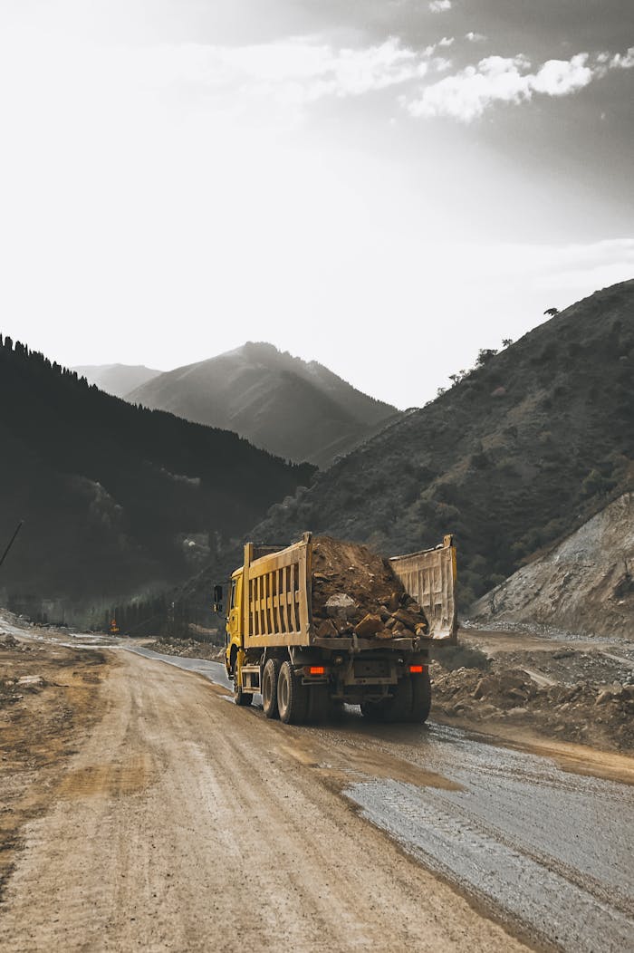 A yellow dump truck carrying dirt on a winding unpaved road through mountainous terrain.